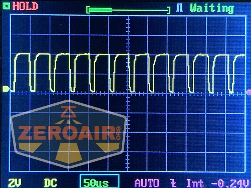 A digital oscilloscope screen displays a repeating square waveform signal. Settings include 2V per division, 50 microseconds per division, and a DC coupling. The word “Waiting” is shown at the top right.