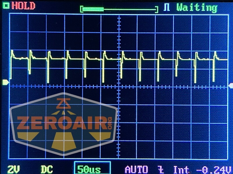 An oscilloscope screen displays a repeating square waveform pattern. The settings show 2V DC and 50 microseconds per division. The screen includes a “HOLD” status and the text ZEROAIR.org in a yellow and orange overlay.