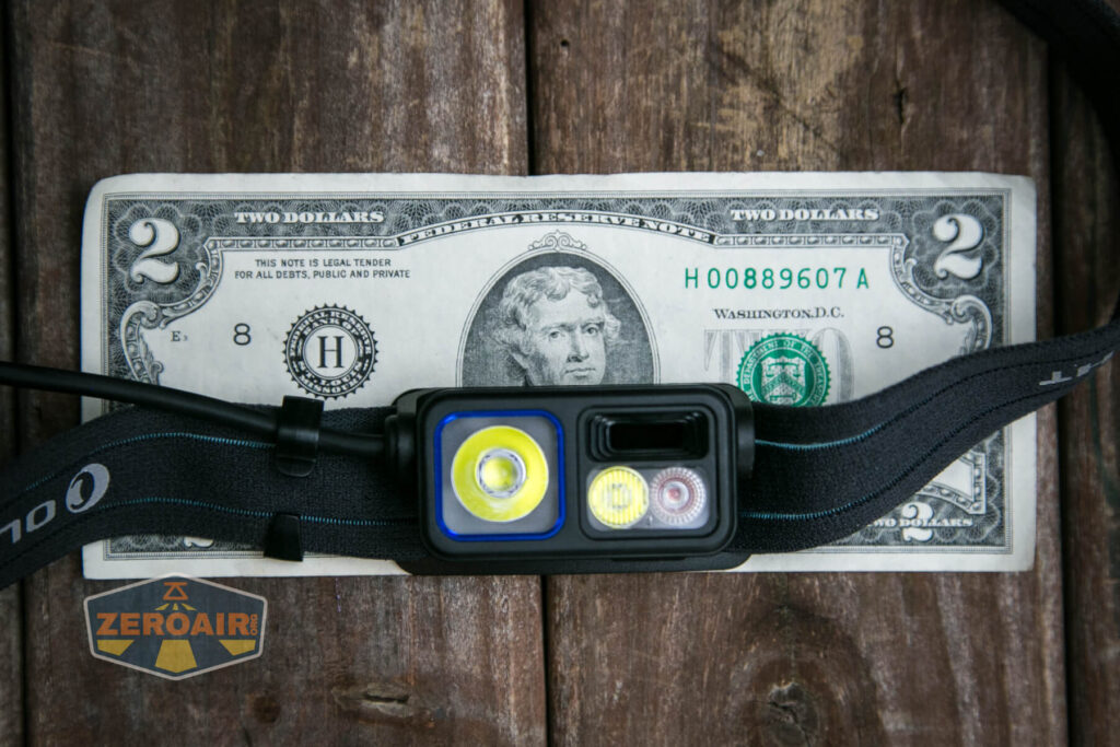 A black headlamp rests on a two-dollar bill atop a wooden surface. A ZeroAir logo is visible in the lower left corner of the image.