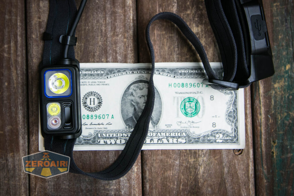 A headlamp with three LED lights and a black strap is placed on top of a two-dollar bill on a wooden surface. The corner of a Zeroair logo sticker is visible in the lower left corner.