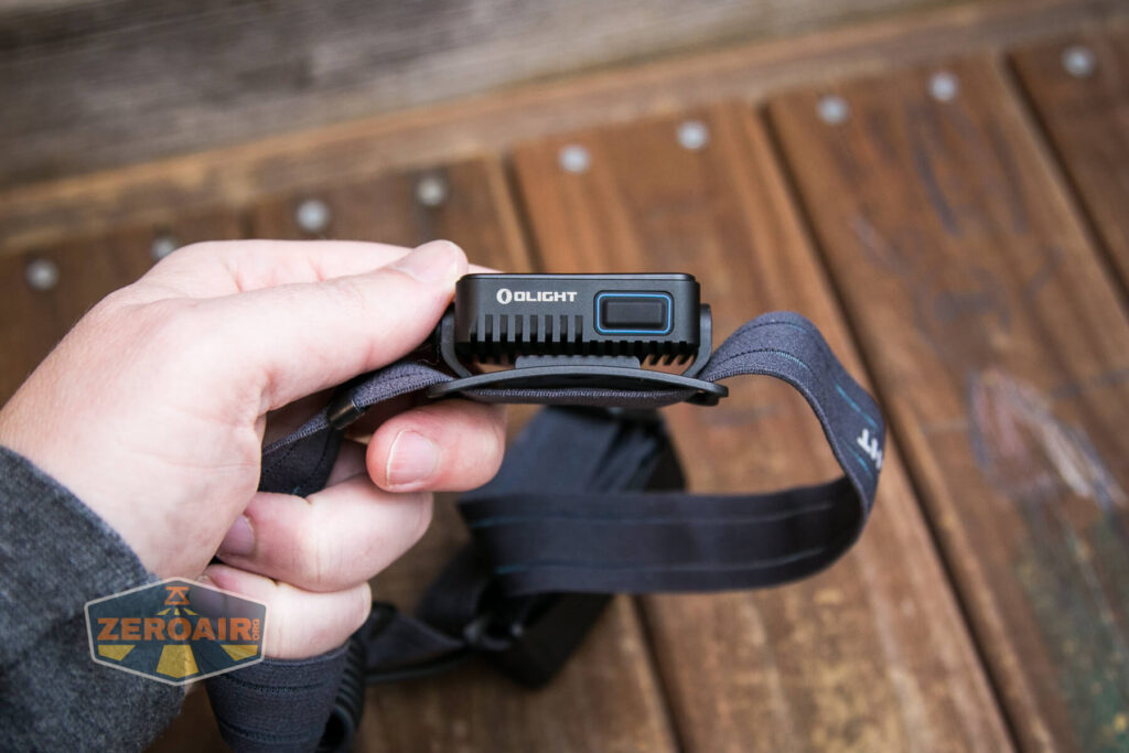 A person holds an Olight-branded black headlamp with an adjustable strap, positioned above a wooden surface with metal screws. The ZeroAir logo is visible in the bottom left corner.