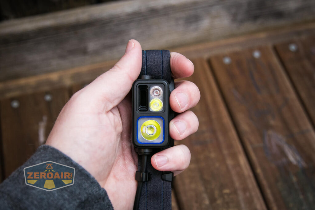 A hand holding a compact black headlamp with three distinct front lenses, attached to a dark elastic strap, against a background of wooden boards.