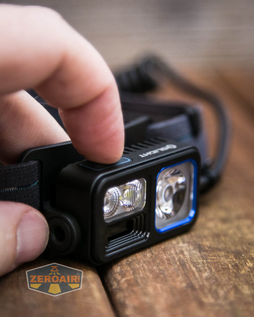 A close-up of a persons fingers about to press the power button on a black headlamp with two lenses. The headlamp is attached to an adjustable strap. A ZeroAir logo is visible in the corner.