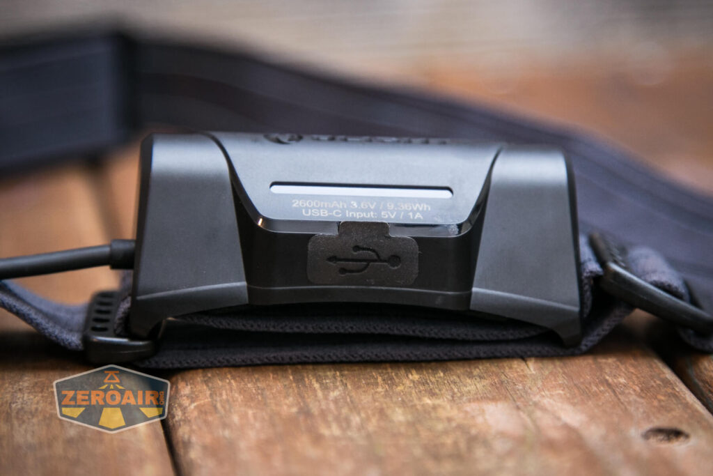 Close-up of a black headlamp with a visible USB-C charging port and specifications label, showing 2600mAh battery and input details, resting on a wooden surface with a ZeroAir logo in the corner.