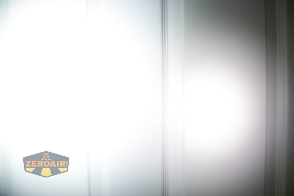 A bright, focused white light beam shines on a white paneled door. The ZeroAir logo is in the bottom left corner. The door details are slightly blurred due to the intensity of the light.