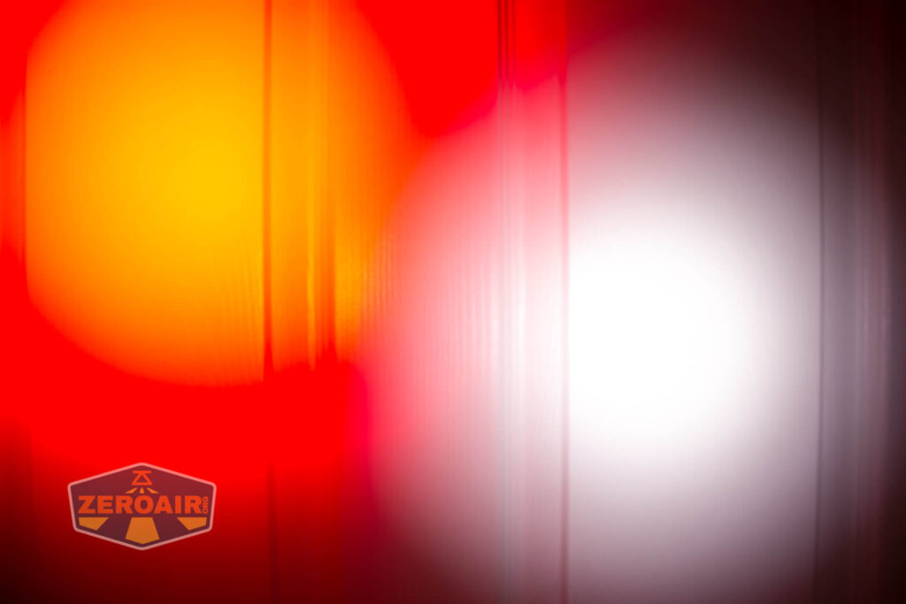 A blurred image with overlapping red, orange, and white circular light spots on a vertically striped background. A logo reading ZEROAIR is visible in the bottom left corner.