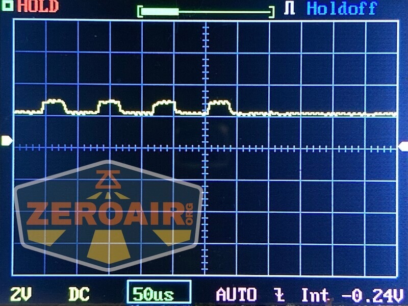 An oscilloscope display shows a yellow waveform with small peaks and valleys, gridlines, and measurement indicators. The screen includes labels such as Holdoff, 2V, 50us, and a transparent ZeroAir logo.