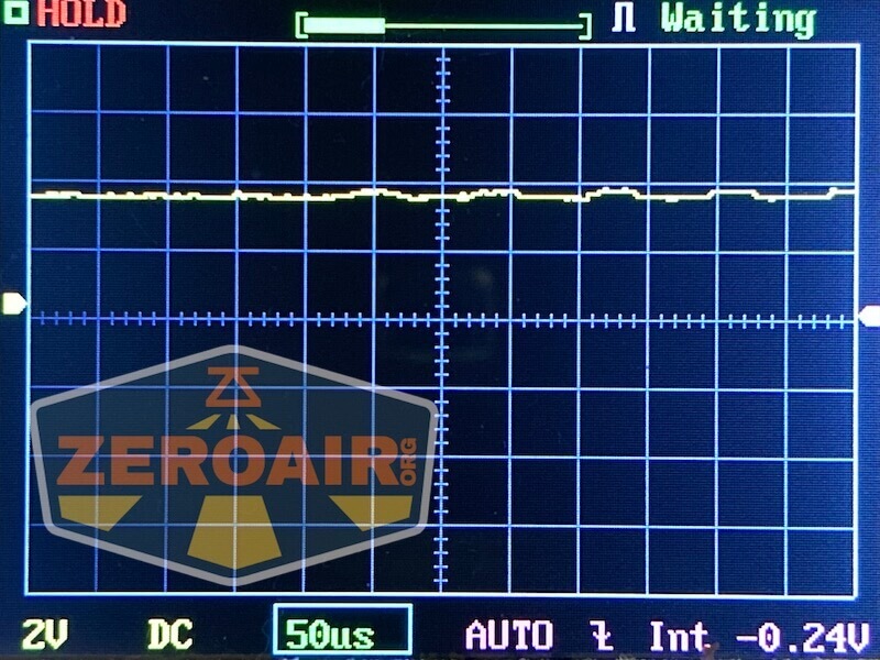 A digital oscilloscope screen displays a mostly flat yellow waveform with minor fluctuations. The screen shows settings such as 2V, DC, 50µs, AUTO, and an average voltage reading of -0.24V. A ZEROAIR.ORG watermark is present.