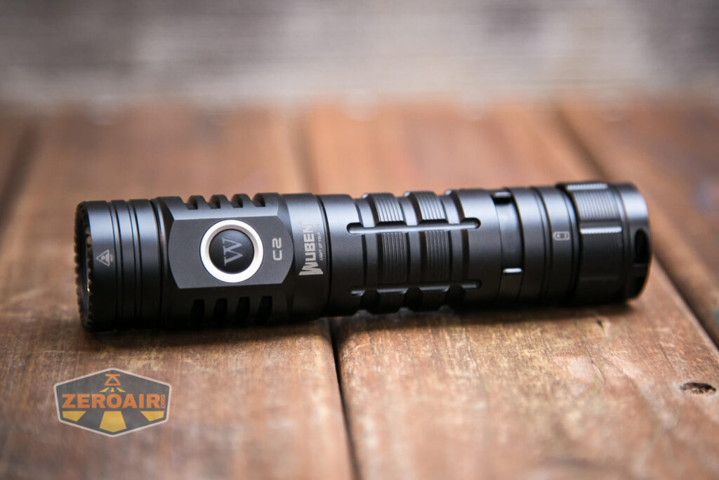 A black, cylindrical Wuben C2 flashlight with a side button lies on a wooden surface. The flashlight features textured grip sections and branding. The ZeroAir logo appears in the bottom left corner.