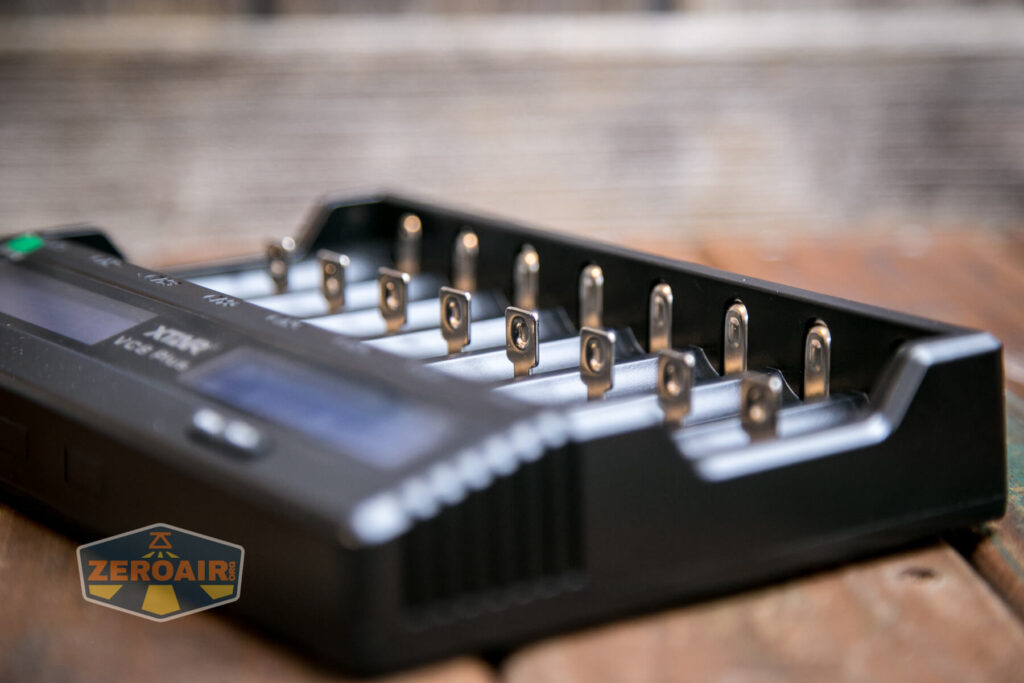 A close-up of a black battery charger with multiple empty charging slots, placed on a wooden surface. The charger displays metal contacts, and a blurred ZEROAIR logo is visible in the foreground.