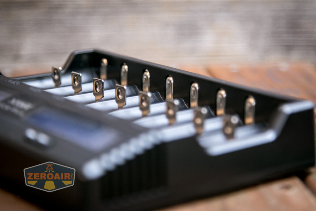 A close-up view of an empty black battery charger with eight slots, placed on a wooden surface. A logo reading ZEROAIR is visible in the bottom left corner of the image.