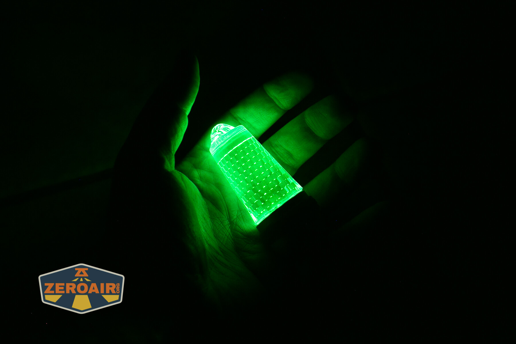A glowing green keychain light is held in a persons hand in the dark. The light illuminates the hand, and a ZEROAIR logo appears in the lower left corner of the image.