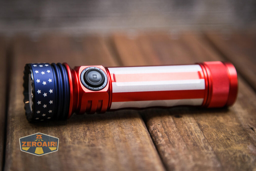 A red, white, and blue flashlight with an American flag design rests on a wooden surface. The flashlight features a prominent side button and a star pattern near the lens. The ZeroAir logo appears in the bottom left corner.