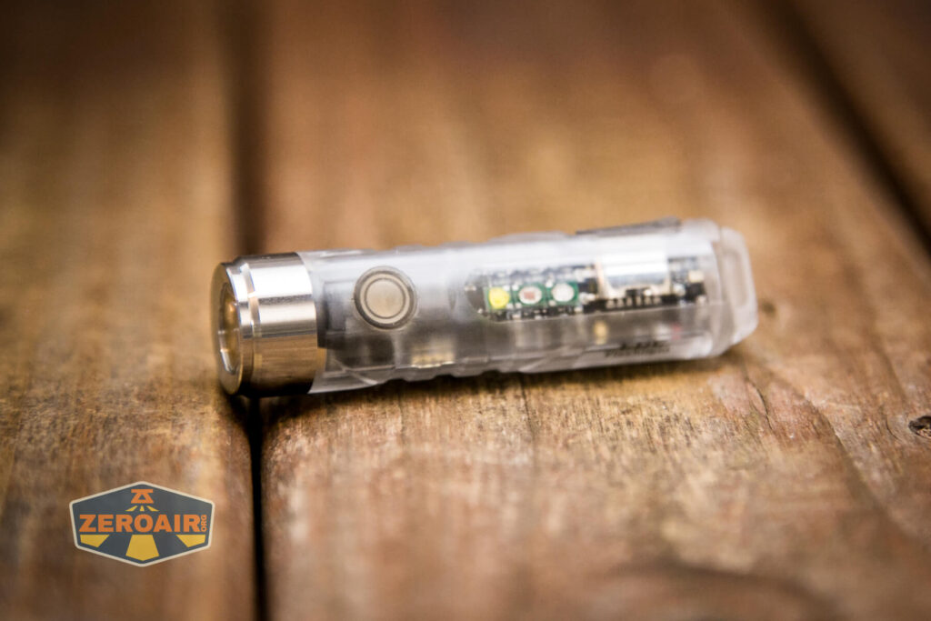 A small, transparent flashlight with visible internal components lies on a wooden surface. The flashlight has a metal button and ZeroAir branding in the corner of the image.