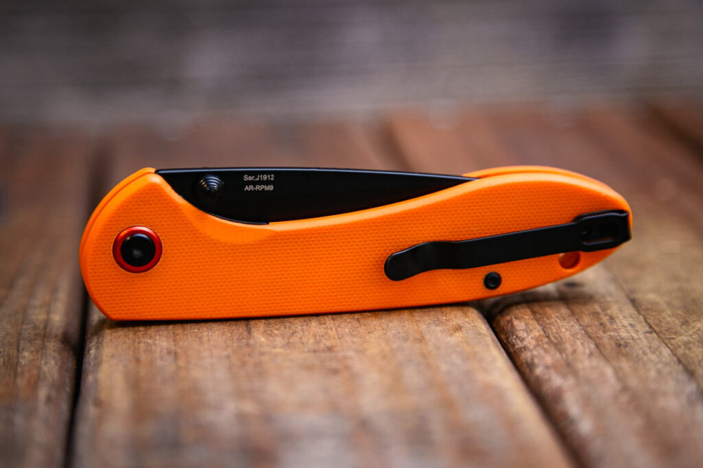 A closed folding pocket knife with an orange handle and black clip is resting on a wooden surface. The blade is partially visible, and there is a red accent near the hinge.