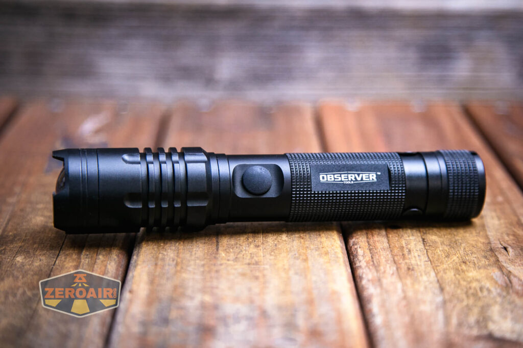 A black Observer FL1000 Zoomie flashlight labeled OBSERVER lies on a wooden surface. The flashlight has a textured grip and a button on its side. The ZeroAir logo is visible in the lower left corner of the image.