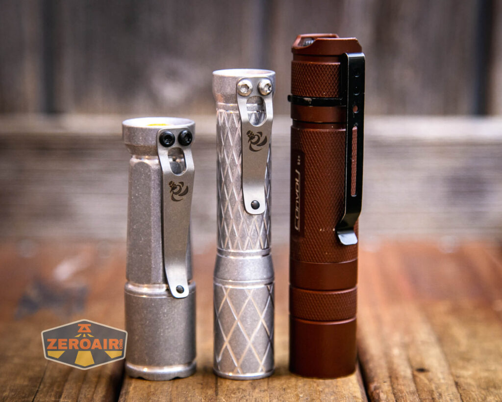 Three flashlights stand upright on a wooden surface. The two on the left are silver with different patterns, while the one on the right is brown and slightly taller. A ZeroAir logo is visible in the bottom left corner.