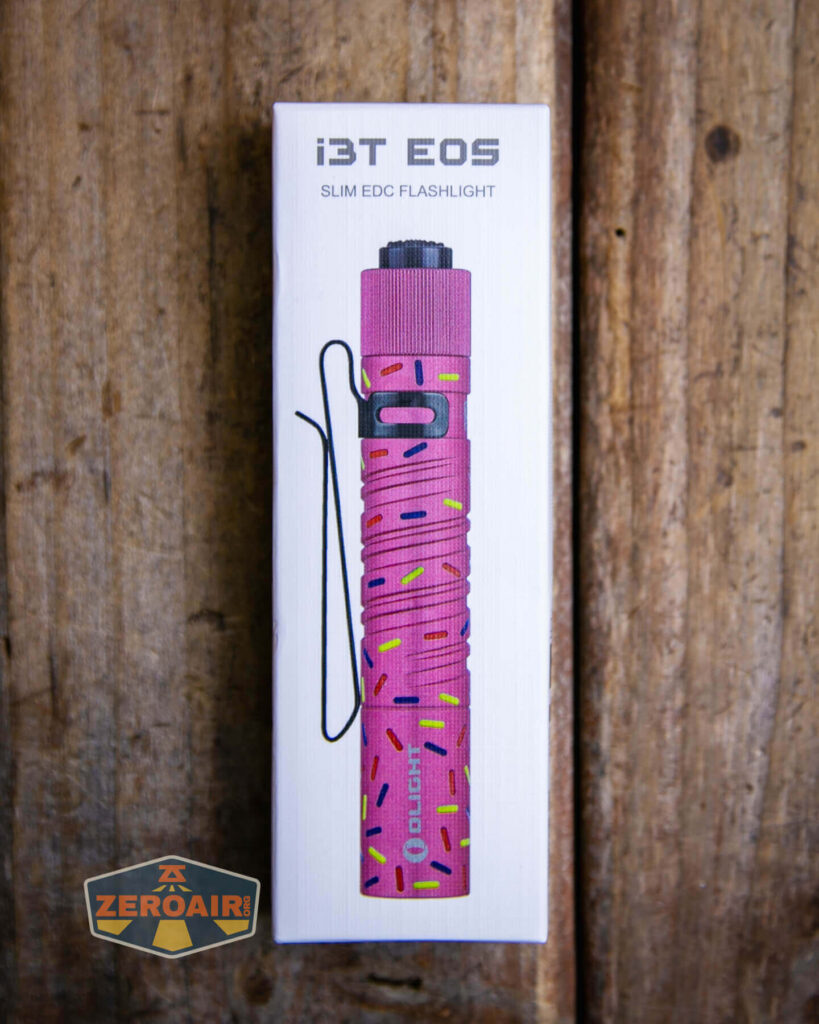 A boxed Olight i3T EOS slim EDC flashlight in a pink color with a sprinkle pattern is shown on a wooden surface. The ZeroAir logo appears in the bottom left corner of the image.