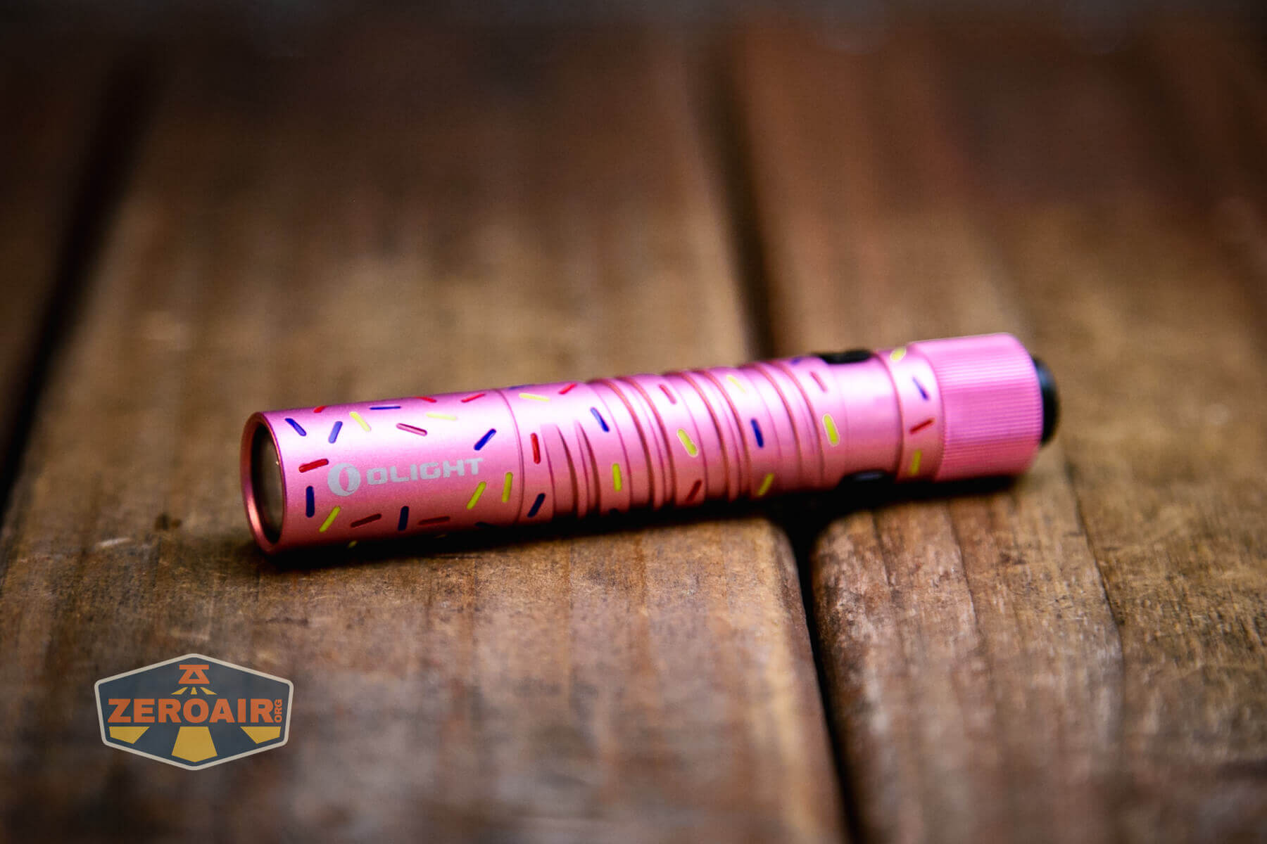 A small, pink Olight i3T EOS Dessert Warrior flashlight with colorful sprinkle-like patterns lies on a wooden surface. The flashlight has the brand name Olight on its side, and a "Zeroair" logo appears in the lower left corner.