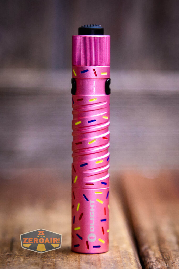 A pink Olight flashlight with a textured grip and colorful sprinkle-like patterns stands upright on a wooden surface. The background is blurred, and a ZeroAir logo is visible in the lower left corner.
