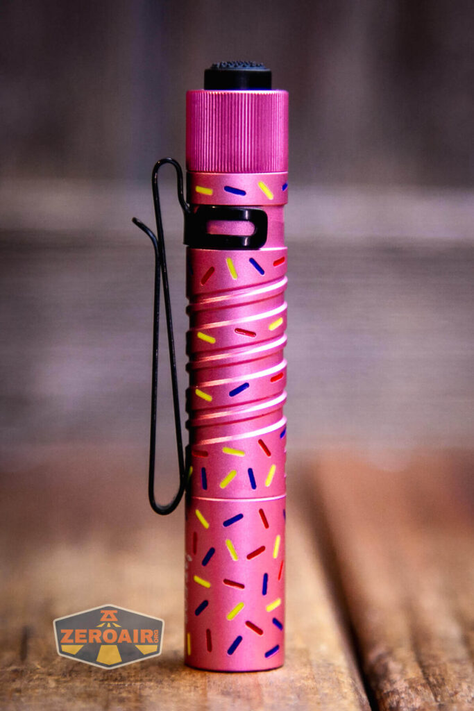 A small, cylindrical pink flashlight stands upright on a wooden surface. It has a black pocket clip and is decorated with colorful sprinkle-like patterns. A ZeroAir logo is in the bottom left corner of the image.