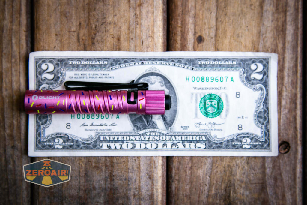 A pink Olight flashlight with a sprinkle pattern rests on a two-dollar bill on a wooden surface. The ZeroAir logo appears in the bottom left corner of the image.