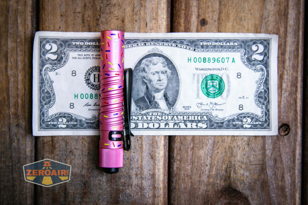 A pink flashlight with a black clip rests horizontally on a two-dollar US bill, both placed on a wooden surface. A ZeroAir logo sticker is visible in the lower left corner of the image.