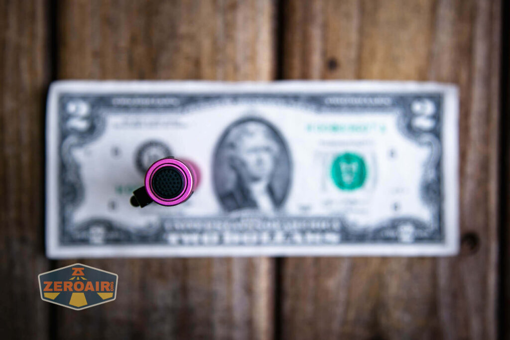 A small, pink cylindrical object stands upright on a two-dollar bill placed on a wooden surface. The bills design and logo reading ZEROAIR are visible in the image.