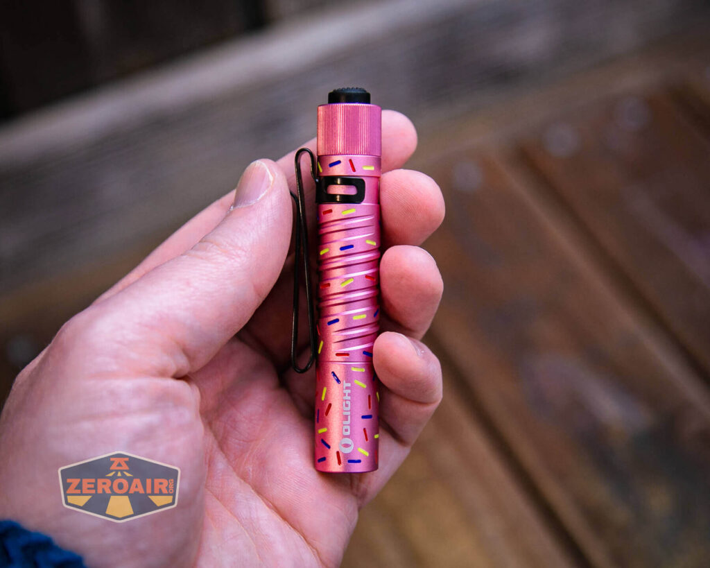 A hand holds a small pink flashlight with a spiral pattern and a black pocket clip. The flashlight is held over a wooden surface, and a ZeroAir logo appears in the lower left corner.