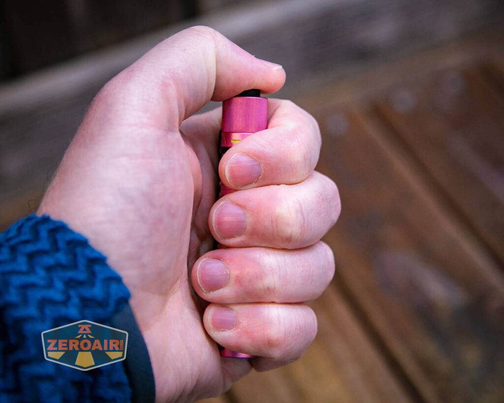 A person’s hand grips a small, cylindrical Olight i3T EOS Dessert Warrior flashlight with their thumb pressing the black button on top; a wooden surface is visible in the background.