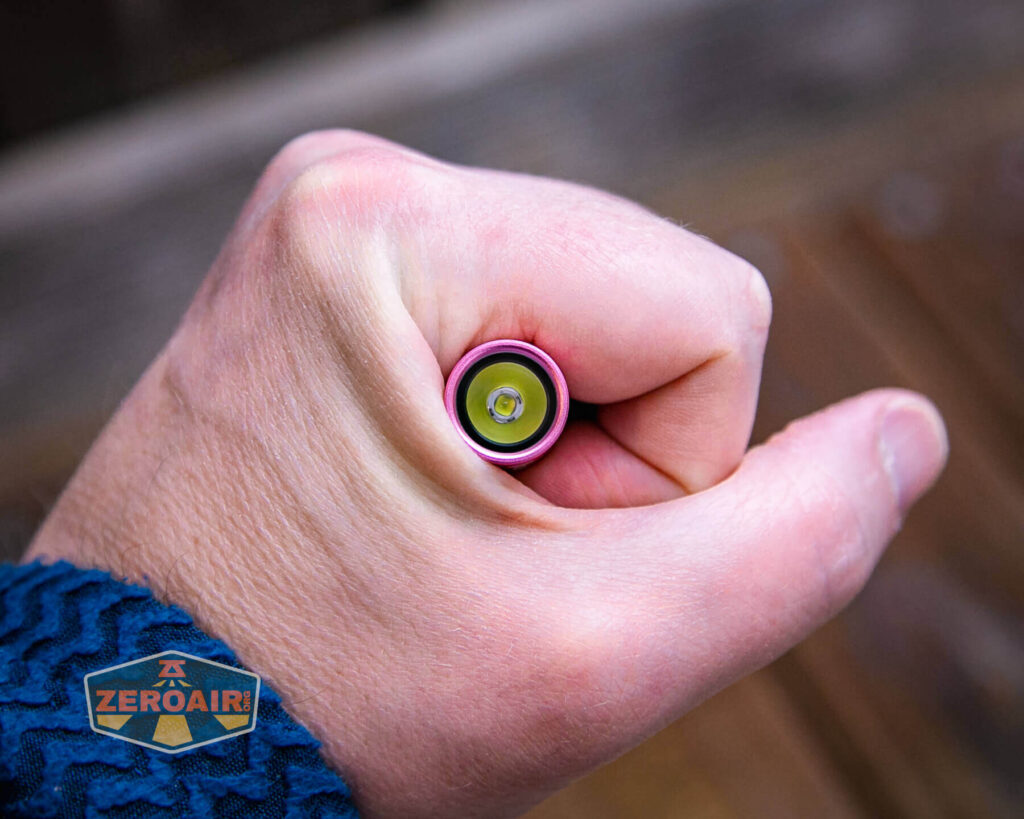 A hand holding a cylindrical object with a visible green and silver circular component at one end, viewed from above. The persons sleeve is blue, and there is a ZeroAir logo in the bottom left corner.