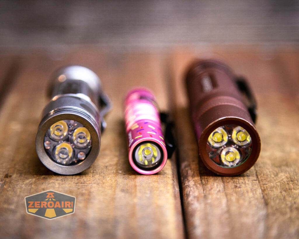 Three LED flashlights, each with three bulbs, are standing upright on a wooden surface. The flashlights are silver, pink, and brown. A small logo in the lower left corner reads ZEROAIR. The background is blurred.