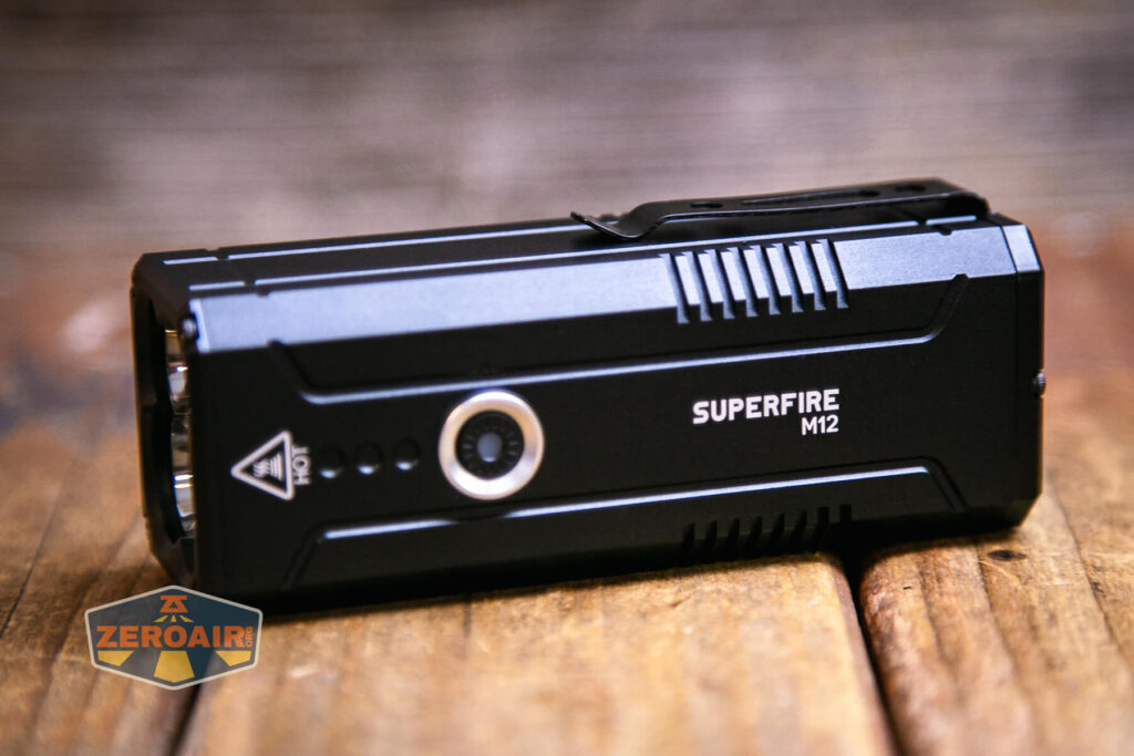 A black Superfire M12 flashlight lies on a wooden surface, angled to show its side and top. The brand and model name are printed on the side, and a round button is visible. The ZeroAir logo appears in the bottom left corner.