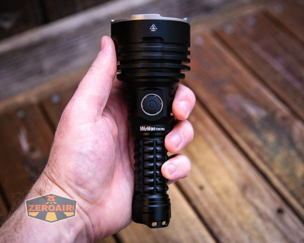 A hand holds a large black Wurkkos TS32 flashlight with a ribbed grip and prominent power button. The background is a wooden surface, and a ZeroAir logo is visible in the lower left corner.