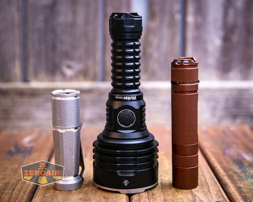 Three flashlights of varying sizes and colors are standing upright on a wooden surface with a blurred wooden background. The largest flashlight is black, the smallest is silver, and the medium-sized one is brown.