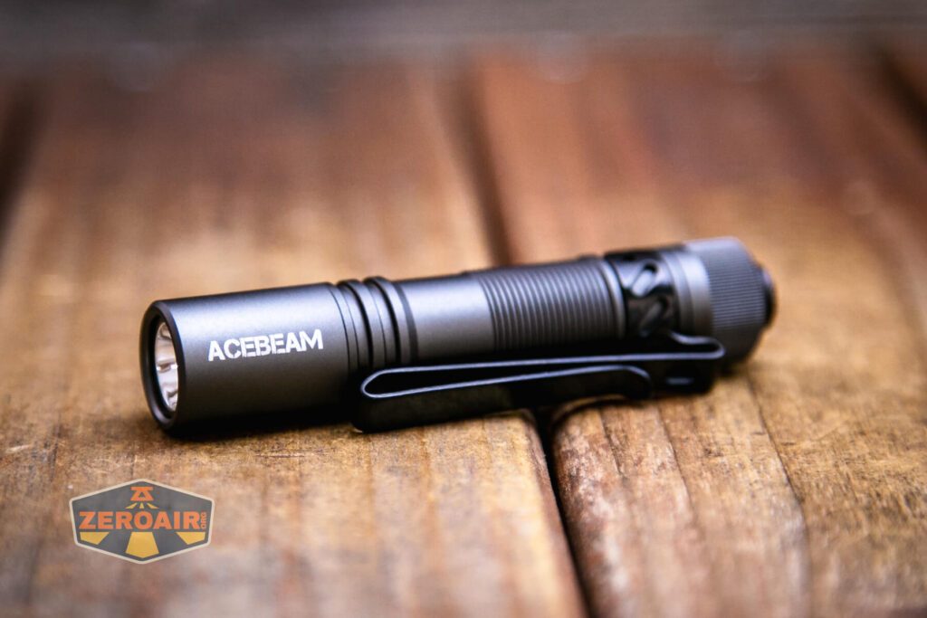 A black Acebeam Pokelit AA Gray flashlight with a pocket clip rests on a wooden surface. The flashlight is positioned horizontally, and the ZeroAir logo is visible in the lower left corner.