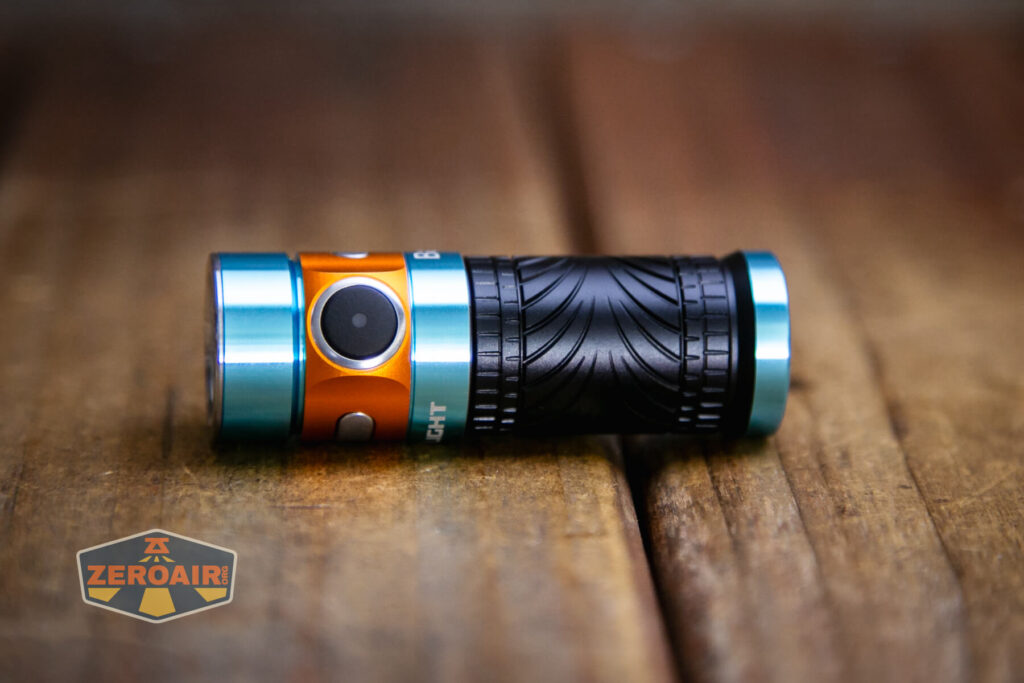 A compact flashlight with blue and orange metallic accents and a textured black grip is lying sideways on a wooden surface. The ZeroAir logo is visible in the bottom left corner.