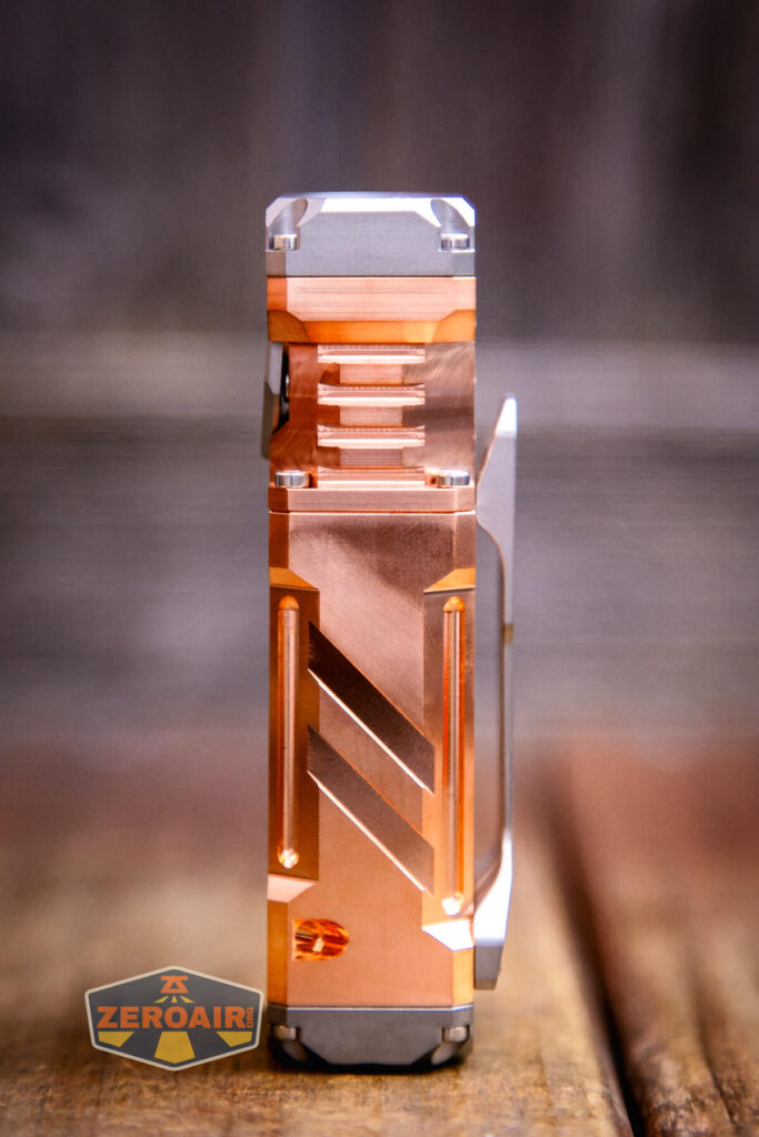 A metallic flashlight with a copper finish, geometric patterns, and a side clip stands upright on a wooden surface with a blurred background; a small ZeroAir logo is in the lower left corner.
