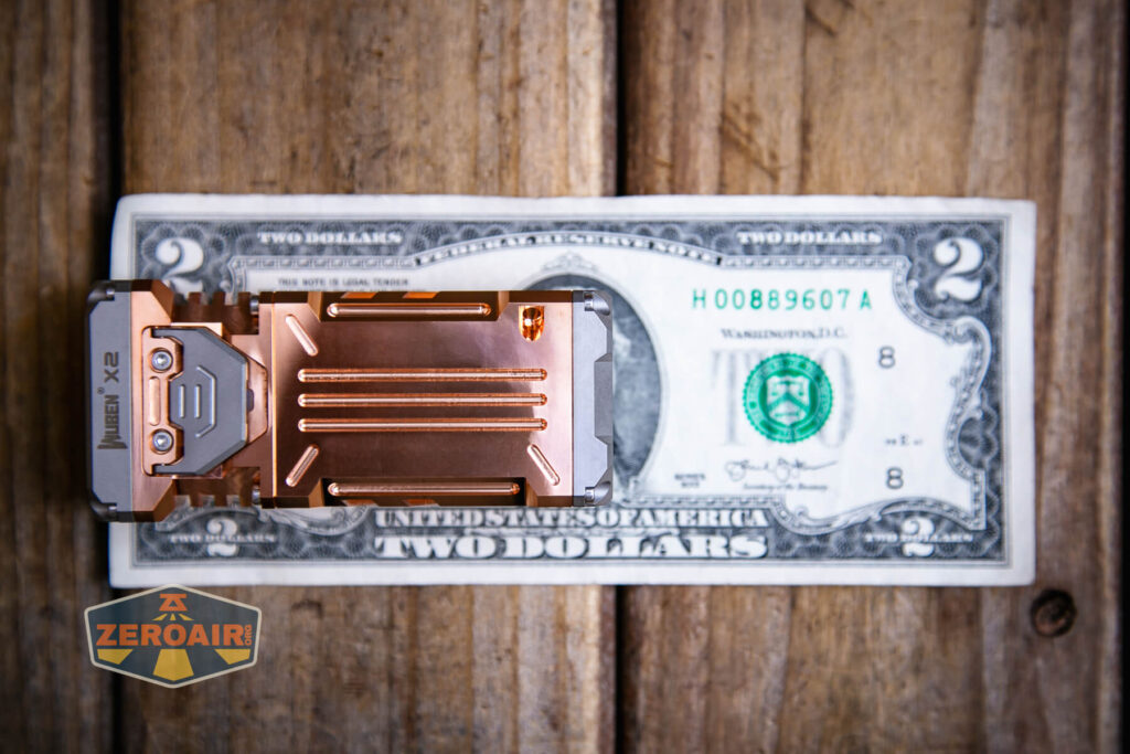 A metallic, copper-colored object labeled Imalent is placed on top of a U.S. two-dollar bill on a wooden surface. A ZeroAir sticker is visible in the lower left corner.