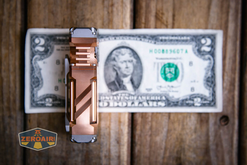 A metal flashlight with a grooved body is placed vertically on top of a U.S. two-dollar bill on a wooden surface. A ZeroAir logo is visible in the lower left corner of the image.