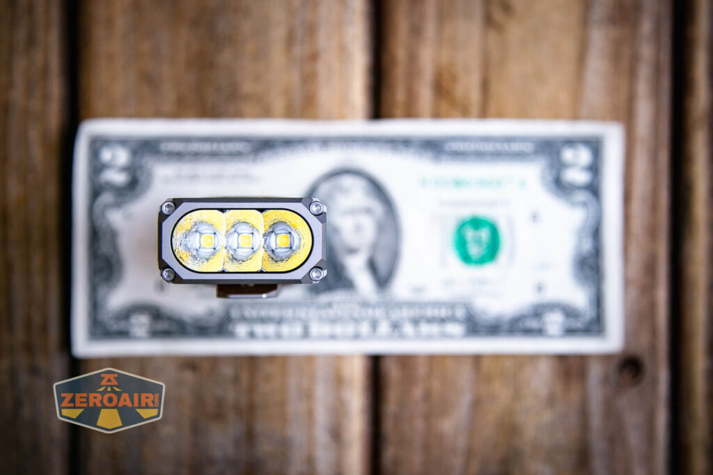 A compact flashlight with three LED bulbs is placed vertically above a two dollar bill on a wooden surface. A ZeroAir logo appears in the bottom left corner.