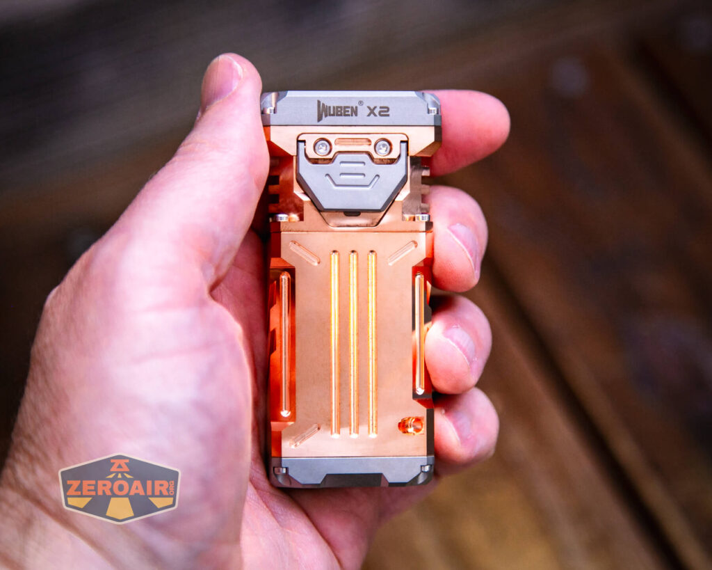 A hand holding a rectangular, metallic orange and gray flashlight labeled “Wuben X2.” The background is blurred, and there is a ZeroAir logo in the lower left corner.