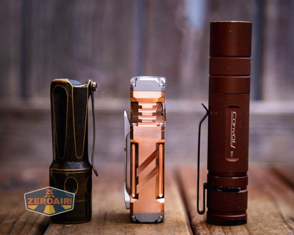 Three flashlights with different designs and finishes, including brass, copper, and matte brown, stand upright on a wooden surface against a blurred wood background. A ZeroAir logo appears in the lower left corner.