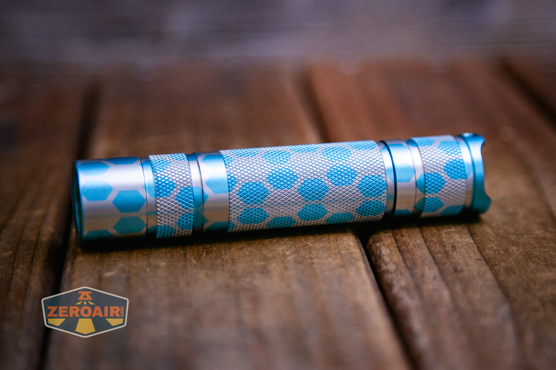 A small, cylindrical Honeycomb Engraved Convoy S2+ flashlight with a blue and silver hexagon pattern lies horizontally on a wooden surface. A logo reading ZeroAir is visible in the lower left corner.