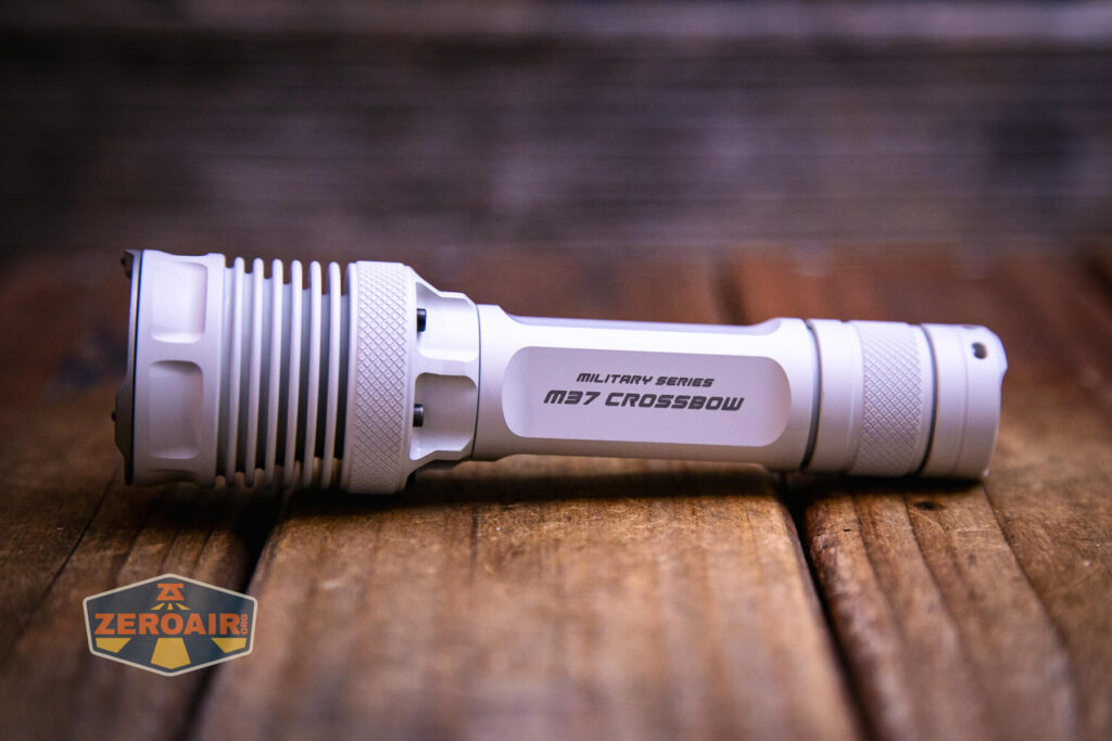 A white JETBeam M37 Crossbow flashlight labeled “M37 CROSSBOW Military Series” lies on a wooden surface with a blurred wooden background. The ZeroAir logo is in the bottom left corner of the image.