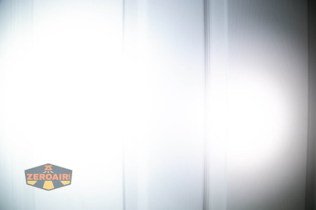 A white vertical surface illuminated by a bright flashlight beam, creating a strong glare and shadows. The ZeroAir logo is visible in the lower left corner of the image.