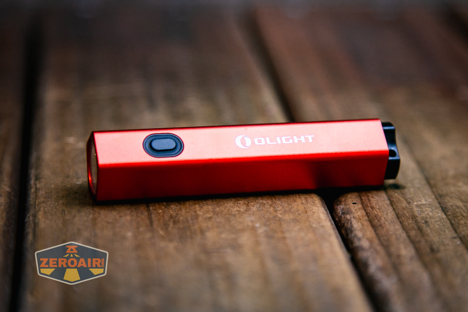 A red Olight Diffuse EDC Pocket Flashlight with a black button and lens lies on a wooden surface. A small ZeroAir logo is visible in the lower left corner.