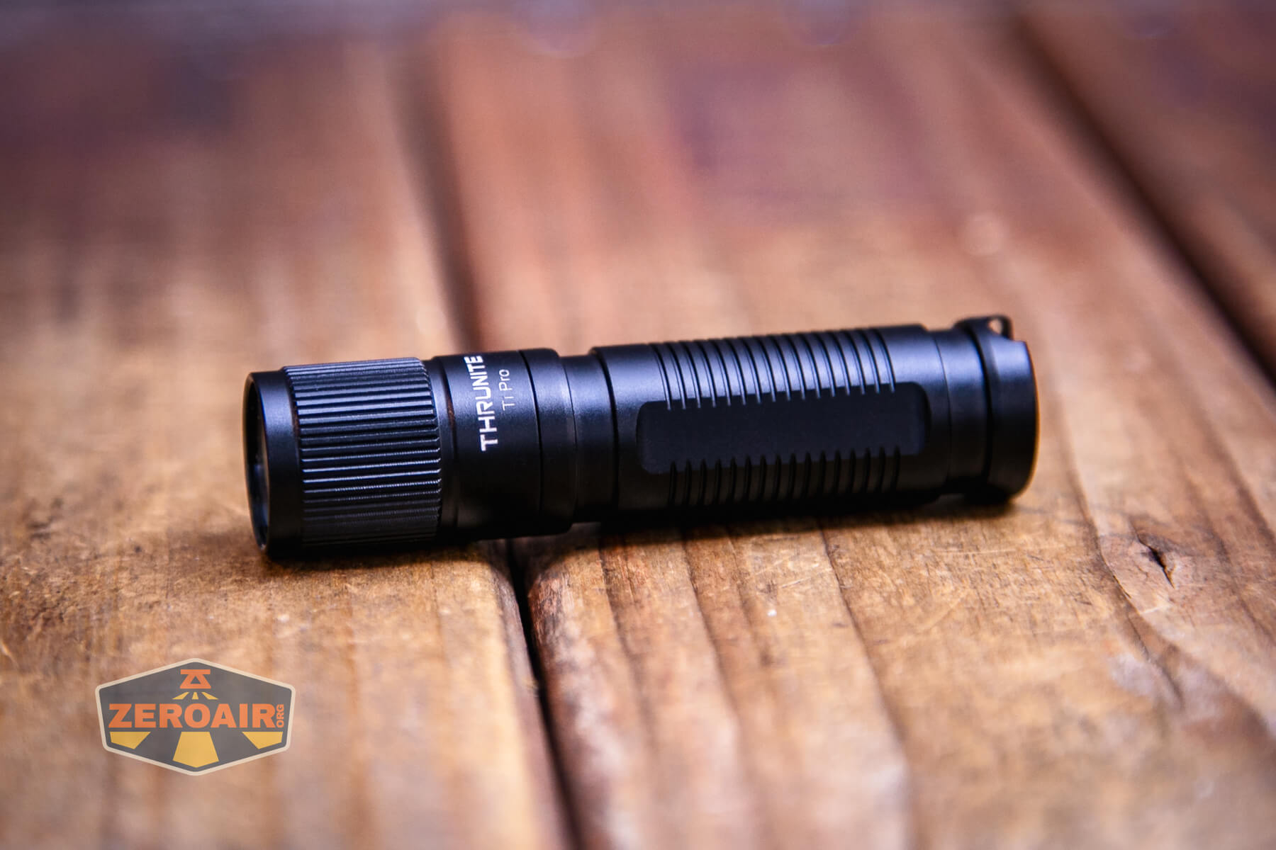 A small black ThruNite T1 flashlight lies on a wooden surface, showcasing its textured grip and ridged bezel. Next to it, the ThruNite Ti Pro flashlight rests nearby. The ZeroAir logo is visible in the bottom left corner of the image.