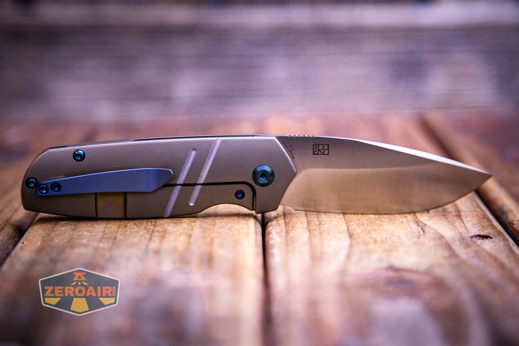 A Kansept Turaco Titanium Knife with a metallic handle and a partially open blade lies on a wooden surface. The ZeroAir logo is visible in the bottom left corner of the image.