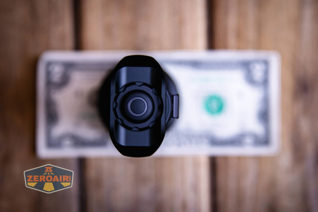 A top-down view of a black flashlight placed upright on a U.S. two-dollar bill, with a wooden surface underneath. A ZeroAir logo appears in the bottom left corner of the image.